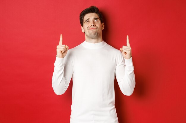 Concept of winter holidays doubtful handsome man in white sweater looking skeptical and pointing fin...