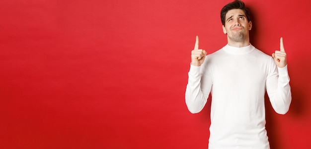 Concept of winter holidays Doubtful handsome man in white sweater looking skeptical and pointing fingers up at logo standing against red background