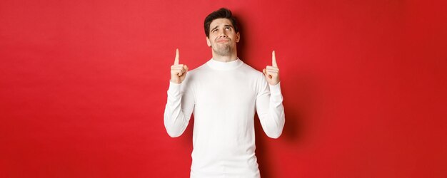 Concept of winter holidays. Doubtful handsome man in white sweater, looking skeptical and pointing fingers up at logo, standing against red background.