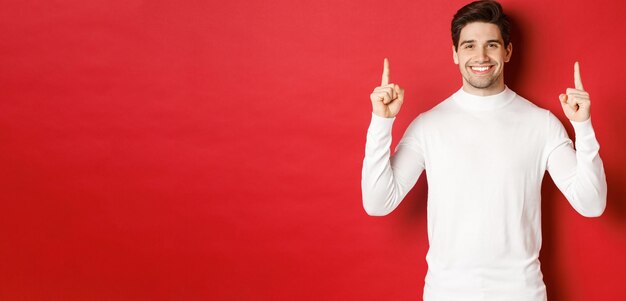 Concept of winter holidays Goodlooking young man with bristle wearing white sweater showing christmas advertisement on copy space pointing fingers up and smiling red background