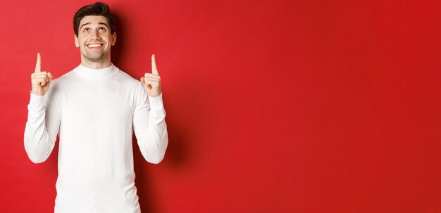 Concept of winter holidays Image of happy attractive man in white sweater smiling while pointing and looking up at copy space on red background