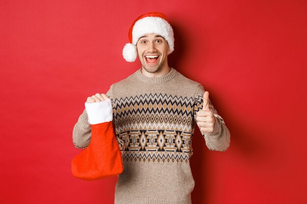 Концепция зимних праздников, нового года и торжества. Веселый красавец в новогодней шапке и свитере, показывает рождественский чулок с конфетами и подарками, показывает палец вверх