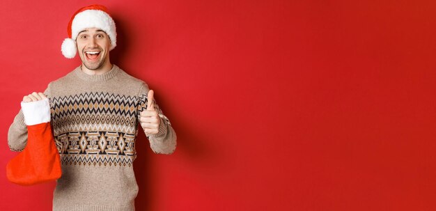 Концепция зимних праздников, нового года и празднования. Веселый красивый мужчина в новогодней шапке и свитере, демонстрирующий рождественский чулок с конфетами и подарками, поднимающий палец вверх.