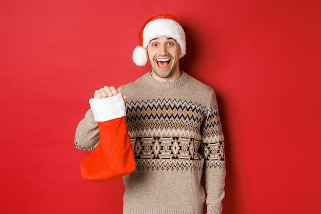 Concept of winter holidays, new year and celebration. Cheerful and surprised adult man receiving candies on Saint Nicholas day in red stocking, standing amazed over red background.