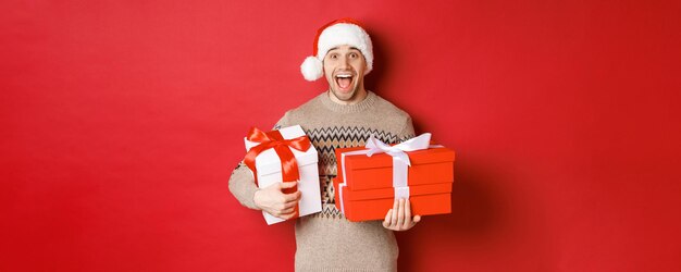 Concept of winter holidays, new year and celebration. Image of amazed and happy man in sweater and santa hat, holding christmas gifts and shouting for joy, red background