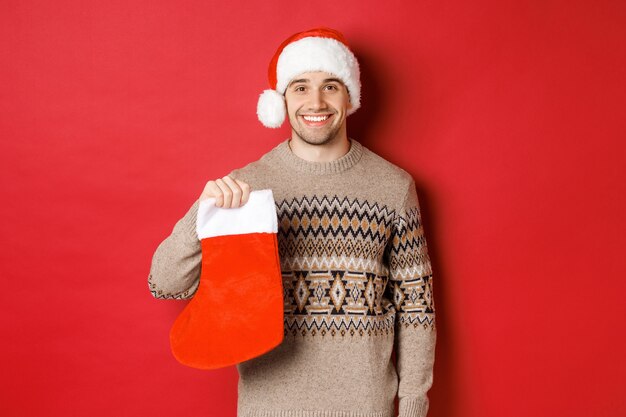 Concept of winter holidays, new year and celebration. Image of handsome smiling man in santa hat and sweater, holding christmas stocking for presents and candies, standing over red background