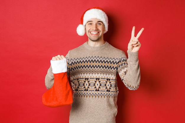 겨울 방학, 새해 및 축하의 개념. 산타 모자와 스웨터를 입은 행복한 미소 짓는 남자의 이미지, 평화 표지와 선물이 든 크리스마스 스타킹 가방, 빨간색 배경