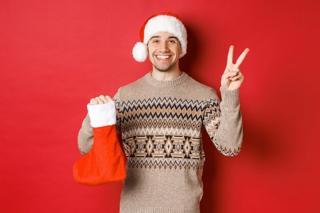 Concept of winter holidays, new year and celebration. Image of happy smiling man in santa hat and sweater, showing peace sign and a christmas stocking bag with gifts, red background