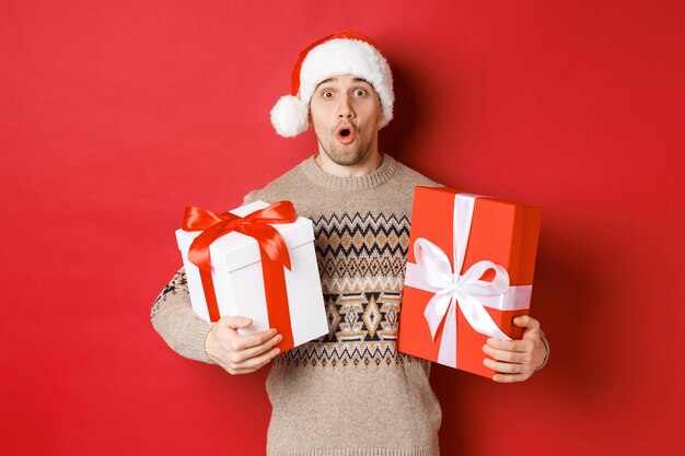 겨울 방학, 새해 및 축하의 개념. 산타 모자와 크리스마스 스웨터를 입은 놀란 매력적인 남자의 이미지, 선물을 받고, 선물을 들고 놀란 표정