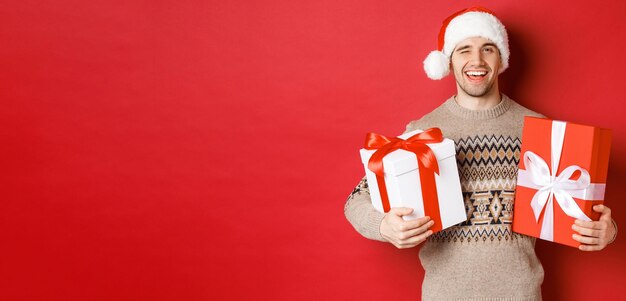 Concept of winter holidays new year and celebration Portrait of confident and cheeky young man prepared gifts for christmas winking and holding presents standing over red background