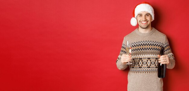 Concept of winter holidays new year and celebration Portrait of handsome man in santa hat and sweater holding champagne and making toast on christmas party standing over red background