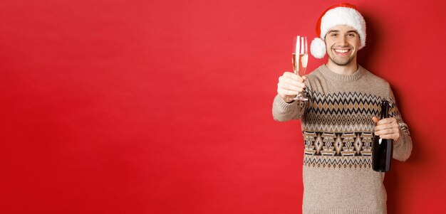 Concept of winter holidays new year and celebration Portrait of handsome man in santa hat and sweater holding champagne raising glass and saying cheers on christmas party