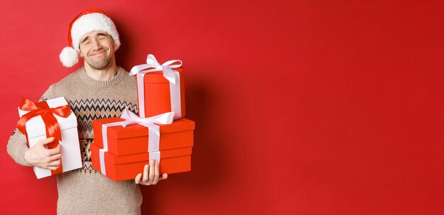Concept of winter holidays, new year and celebration. Portrait of lovely smiling man receiving pile of presents, holding gifts and being touched with surprise, standing over red background grateful