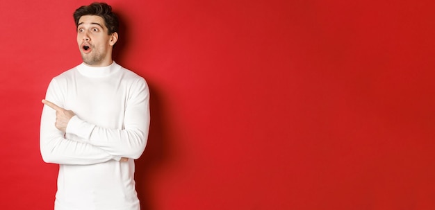 Concept of winter holidays. Portrait of impressed handsome man in white sweater, pointing and looking left with dropped jaw and amazed expression, standing against red background