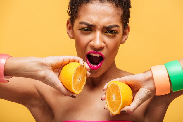 Conceptual amazing afro american woman putting two parts of ripe orange back together on camera isolated, over yellow wall