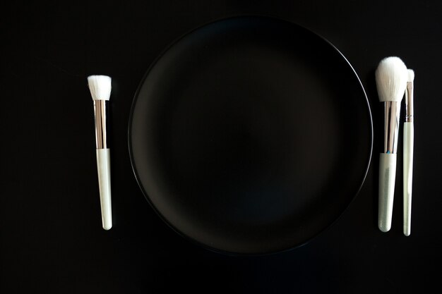 Conceptual image of make up brushes next to dinner plate on black background