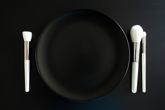 검정색 배경에 있는 저녁 접시 옆에 있는 메이크업 브러쉬의 개념적 이미지