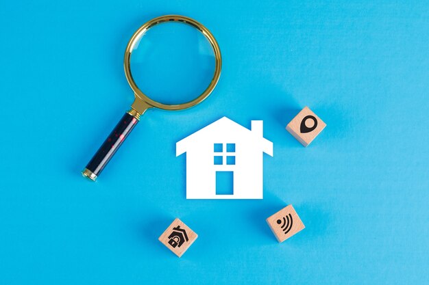 Conceptual of real estate with magnifying glass, wooden blocks, paper home icon on blue table flat lay.