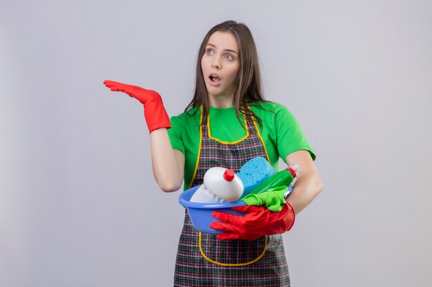 Concered cleaning young woman wearing uniform in red gloves holding cleaning tools raised hand on isolated white wall