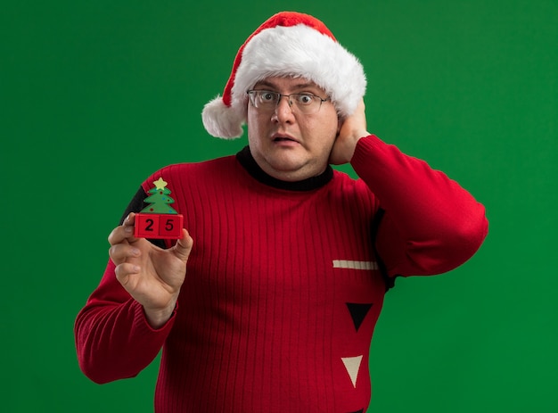 Free photo concerned adult man wearing glasses and santa hat holding christmas tree toy with date looking at camera keeping hand on head isolated on green background