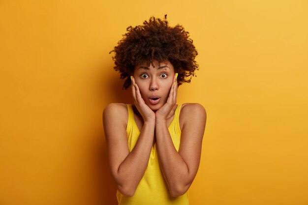 Concerned alarmed curly haired African American woman grabs face, stands shocked and anxious, witnesses terrible accident, stares at something bad, hears scary news, wears bright yellow clothes