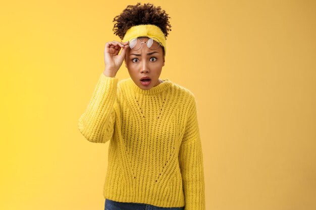 心配している驚いた言葉のないアフリカ系アメリカ人の学生の女の子のドロップジョーテイクオフグラスは目を広げて驚いたドロップアウトされた見た目は信じられないほどのもので、立って質問された欲求不満の黄色の背景。