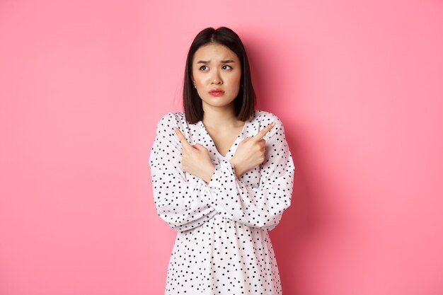 Concerned asian woman having doubts, pointing sideways and looking left with hesitant and sad face, need help with choice, standing over pink background