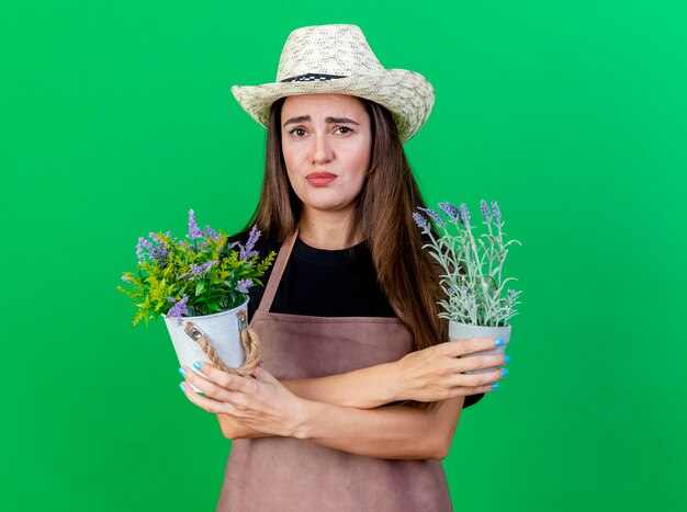 Concerned beautiful gardener girl in uniform wearing gardening hat holding and crossing flowers in flowerpot isolated on green background