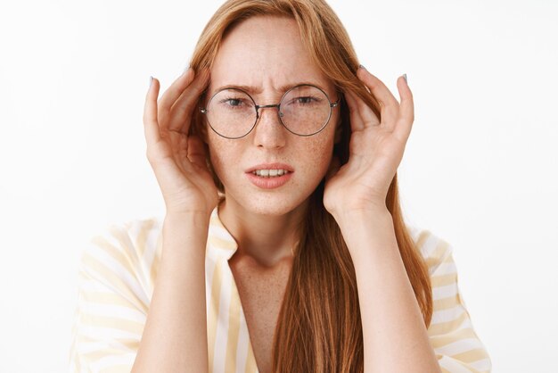 Concerned clueless and perplexed good-looking stylish woman in glasses frowning and squinting holding hands on temples of head being troubled or suffering headache over white wall