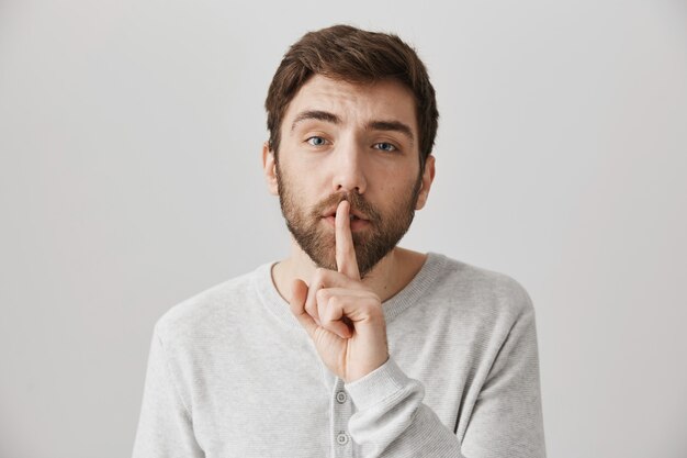 Concerned cute bearded guy show hush sign, say shh to keep voice down