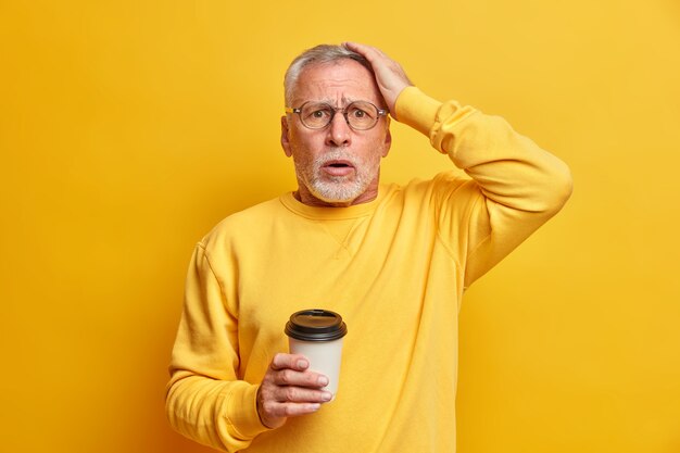 Обеспокоенный смущенный бородатый пожилой мужчина держит руку на голове и ошеломленно смотрит на вход, пьет кофе на вынос, одетый в повседневный джемпер, изолированный над желтой стеной