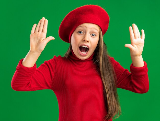 Concerned little blonde girl wearing red beret keeping empty hands in air  isolated on green wall
