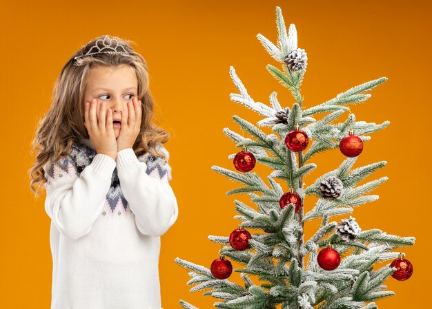 Concerned little girl standing nearby christmas tree wearing tiara with garland on neck covered cheeks with hands isolated on orange background