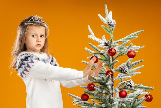 Обеспокоенная маленькая девочка, стоящая рядом с елкой в тиаре с гирляндой на шее, протягивает руки к елке, изолированной на оранжевом фоне