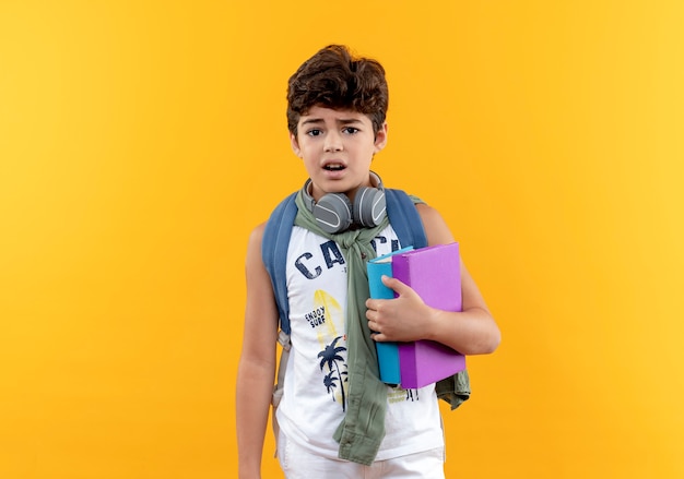 Concerned little school boy wearing back bag and headphones holding books isolated on yellow background