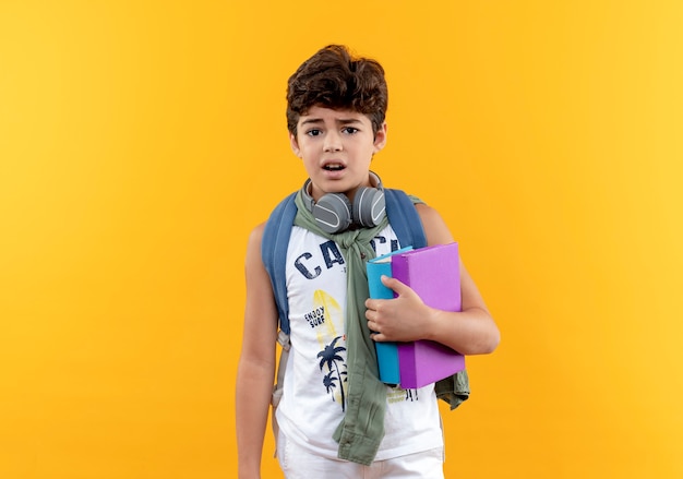 黄色の背景で隔離の本を保持しているバックバッグとヘッドフォンを身に着けている心配の小さな男子生徒