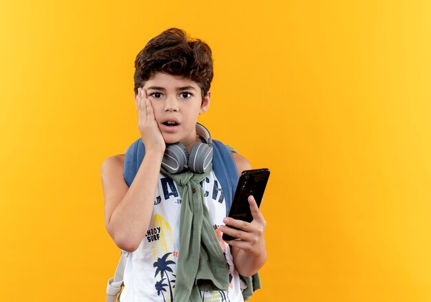 Concerned little school boy wearing back bag and headphones holding phone and putting hand on cheek isolated on yellow background