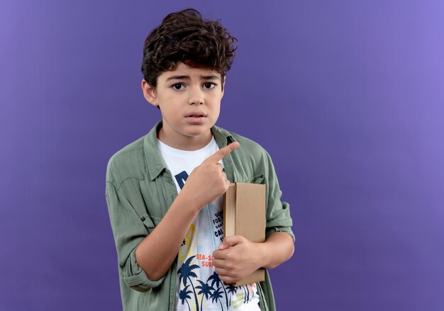 Concerned little schoolboy holding book and points at side isolated on purple wall with copy space