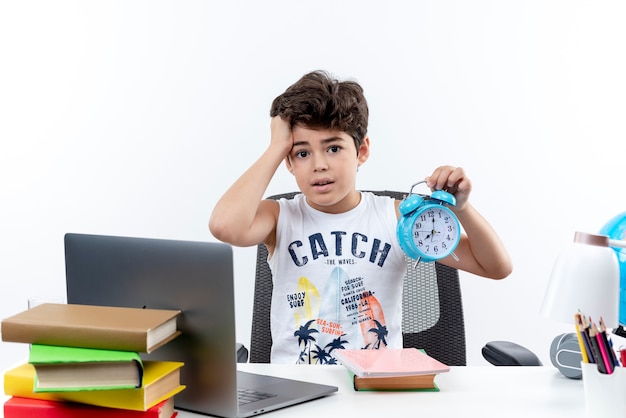 Concerned little schoolboy sitting at desk with school tools holding alarm clock and grabbed head isolated on white background