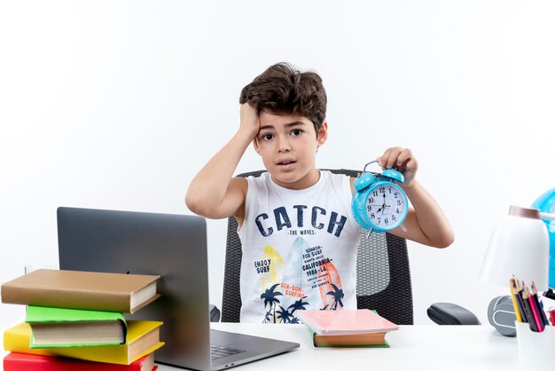 Concerned little schoolboy sitting at desk with school tools holding alarm clock and grabbed head isolated on white background