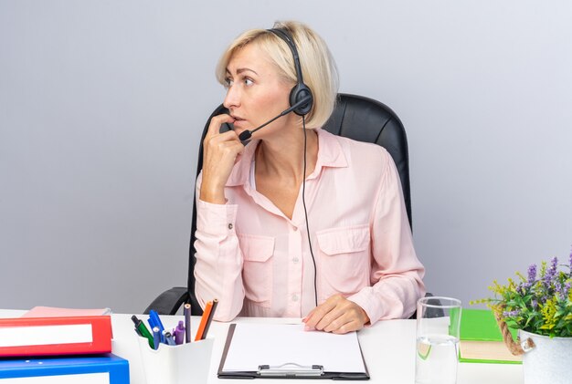 Concerned looking side young female call center operator wearing headset sitting at table with office tools isolated on white wall