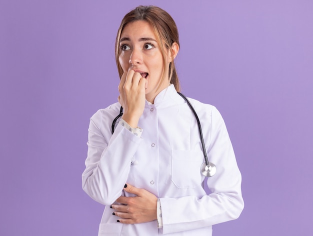 Обеспокоенная смотрящая на бок молодая женщина-врач в медицинском халате со стетоскопом кусает ногти, изолированную на фиолетовой стене