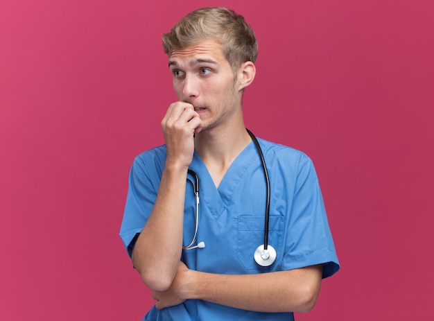 Обеспокоенный глядя на сторону молодого врача-мужчины в униформе врача со стетоскопом кусает ногти, изолированную на розовой стене