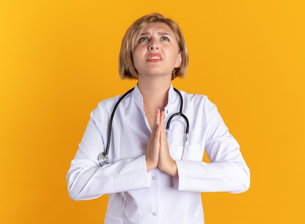 オレンジ色の壁に隔離された祈りのジェスチャーを示す聴診器で医療ローブを着ている若い女性医師を見上げることを懸念