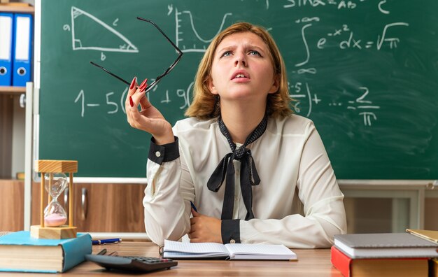 Concerned looking up young female teacher sits at table with school tools holding glasses in classroom