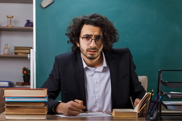 Insegnante maschio interessato con gli occhiali scrive qualcosa su carta seduto al tavolo con gli strumenti della scuola in classe