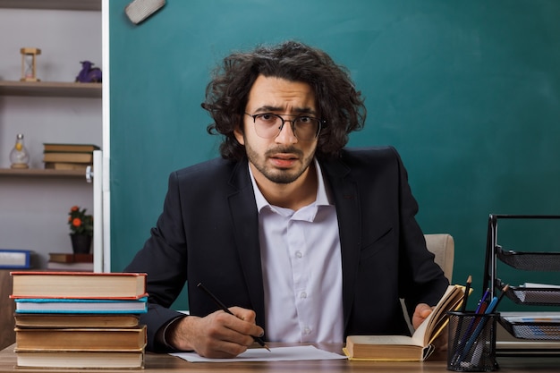 Обеспокоенный учитель-мужчина в очках пишет что-то на бумаге, сидя за столом со школьными принадлежностями в классе