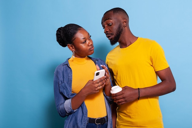 Free photo concerned man spying on girlfriend sms on mobile phone. young woman holding smartphone and browsing internet while her boyfriend looks at notification on screen in studio, expecting betrayal.