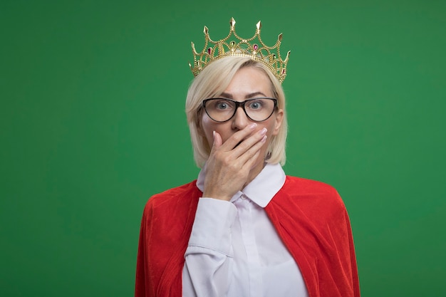 Free photo concerned middle-aged blonde superhero woman in red cape wearing glasses and crown keeping hand on mouth