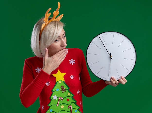 Concerned middle-aged blonde woman wearing christmas reindeer antlers headband and christmas sweater holding and looking at clock keeping hand on mouth isolated on green wall